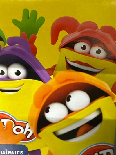 The image displays a close-up of packaging for Play-Doh, featuring brightly colored cartoon characters with large eyes and open mouths. The characters are depicted in a playful and exaggerated style. The dominant colors are yellow, red, orange, and purple. One character, predominantly red, has large, expressive eyes and what appears to be red hair or head decoration. Another character, mostly orange, also has large white eyes with black pupils and a wide mouth. A third character, in purple, is partially visible, with large eyes peeking out. A green hand-like shape is also visible on the left side of the image.
The overall impression is one of fun and creativity, likely associated with a children's toy. The characters' wide eyes and open mouths suggest surprise, excitement, or laughter. The background is a vibrant yellow, reinforcing the cheerful theme.
Partially visible text on the right side of the image includes the stylized "Play-Doh" logo in red and white, with an orange outline. Below this, a blue rectangular label with white text is visible, which reads "couleurs". The "Chociule, Poland" location context suggests this is likely product packaging sold in that region. There are no cues about the time of day or weather. The scene is an indoor product shot, focusing on the product's visual appeal.