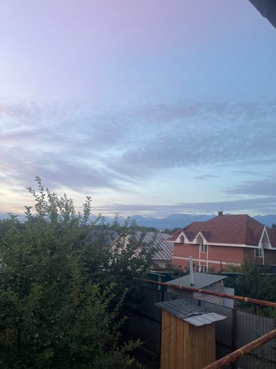 The image depicts an outdoor scene with a focus on the sky and distant mountains. The sky is a soft gradient of purple and blue, with wispy clouds scattered across it. In the middle ground, several residential buildings are visible, characterized by red-tiled roofs and brick exteriors. Some of these houses feature architectural details like dormer windows. A dense cluster of green trees dominates the left foreground, obscuring parts of the buildings. Behind the houses and trees, a range of mountains can be seen in the distance, their peaks lightly dusted with snow, suggesting a cooler climate. The overall impression is one of a suburban or rural setting, possibly during dawn or dusk, given the soft lighting and colors of the sky. The location is identified as Qaskeleñ, Kazakhstan. There are no people or animals visible in the frame. The scene is static, with no discernible actions or interactions taking place.