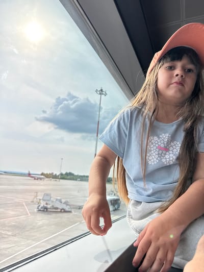 The media file shows a young girl, likely a child, wearing a pink hat and a light blue t-shirt with a floral design. She is positioned in the foreground, looking towards the camera with a neutral expression. Her hair is long and brown, falling around her shoulders. She is seated or leaning near a large window, with her hands visible.
The setting appears to be an airport terminal, viewed from inside through a window. Outside the window, an airport tarmac is visible, with parked aircraft, jet bridges, and airport personnel vehicles. The sky is bright, suggesting it is daytime, and there are some clouds, indicating potentially mixed weather. The sun is visible, casting a bright glare on the window. The reflection of the sky and some of the interior of the airport is visible on the glass, creating a layered effect.
There is no visible text in the image. The overall mood is casual and possibly anticipatory, given the airport setting. The girl's gaze is direct, engaging the viewer. The image is taken from a slightly angled perspective, with the girl appearing to be above the viewer.