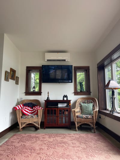 The image shows a cozy living room with a rustic charm, possibly located in Brantingham, United States. The room features two wicker chairs, one adorned with an American flag draped over it, and the other with a light green throw pillow and a floral-patterned cushion. Between the chairs, a wooden cabinet with glass doors holds media equipment. A flat-screen television is mounted on the wall above the cabinet, displaying a scene from "Anne Rice's The Vampire Lestat."
To the left of the television, a small collection of framed pictures hangs on the white wall. To the right, a large window offers a view of lush greenery, and a lamp with a patterned shade sits on the windowsill. Another smaller window with a dark wooden frame is situated to the left of the television, and it appears to be a decorative niche with a lantern and a green pitcher placed inside. The room is illuminated by natural light streaming in from the windows, creating a warm and inviting atmosphere. A pink rug with an intricate floral pattern covers the floor in the foreground. The overall ambiance is one of comfort and relaxation, with a touch of patriotic decoration.