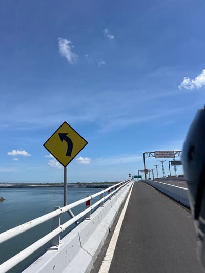 A road on a bridge over water with a bright blue sky and some clouds. A yellow diamond-shaped sign with a black arrow pointing left is on the left side of the road. There are white railings and a concrete barrier along the edge of the bridge. In the distance, there are more bridges or structures. A black object, likely part of a vehicle, is in the foreground on the right. A sign above the road in the distance has red text. This is a view from a moving vehicle, likely a motorcycle, traveling on the Jagatnatha Bridge in Denpasar, Bali, Indonesia, a prominent infrastructure project connecting the Sanur area to the island of Nusa Lembongan.