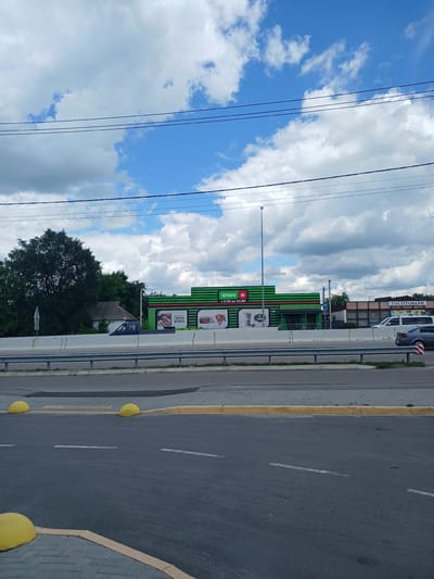 The image depicts a street scene in Hrebinky, Ukraine, with a prominent green and red building featuring the "ФОРА" logo, indicating a supermarket or convenience store. The building is situated behind a concrete barrier and a metal fence. Several vehicles are visible, including a blue pickup truck parked near the building, a white van, and a gray car on the road. The sky is partly cloudy, with a mix of blue sky and white clouds. Power lines are visible stretching across the top of the frame. In the foreground, there is a paved road with yellow bollards and yellow and white road markings. Adjacent to the road is a pedestrian walkway with gray paving stones. Another building with signage reading "ГОСП ТОВАРИ" (Household Goods) is visible to the right of the "ФОРА" building.