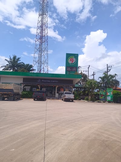 Captured in Plai Phraya, Thailand, the image presents a daytime view of a gas station, identifiable by the prominent green "PT" logo. The station's building houses a "PUNTHAI COFFEE" shop, with vehicles including a pickup truck and a dump truck parked in front. A tall communications tower stands nearby. Additional details include advertising banners, utility poles with power lines, and palm trees, all under a partly cloudy sky.