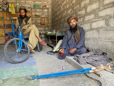 The image depicts two men in a small, brick-walled building in Panjwai, Afghanistan. The building appears to be a shop with shelves stocked with goods. One man, wearing a traditional Afghan hat and clothing, sits in a blue, three-wheeled bicycle, partially disassembled and with one wheel removed. The other man, also wearing traditional Afghan clothing, sits on the floor and has a prosthetic leg. He is holding a rosary in his right hand. Both men appear to be middle-aged. The floor is covered in a brown, patterned rug. There is a pair of crutches lying on the floor to the right of the seated man. The men are not interacting with each other. The scene is lit by natural light coming through a doorway.