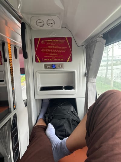 Inside a vehicle, possibly a sleeper bus, a person wearing brown pants and light blue socks rests their feet in a storage compartment. A black backpack is also placed in the compartment.  A panel above the compartment features a white control panel with ventilation controls and a maroon sign with gold lettering indicating "THANH TRUNG TRAVEL" and a hotline number.  A gray window shade is visible above the panel, and part of a window is visible through the shade.  A small pull-out table is visible underneath the storage compartment.