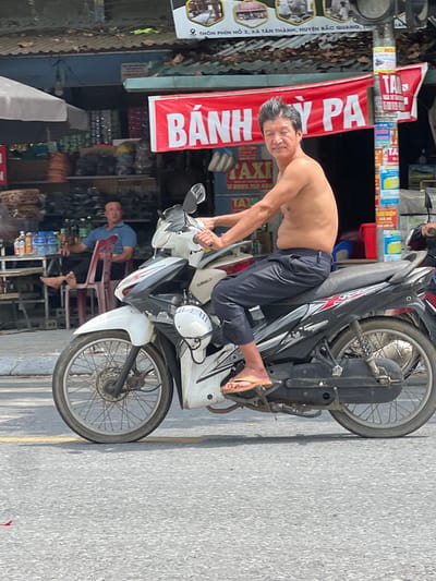 A shirtless man with slicked-back black hair rides a white and black motorcycle down a paved road in Tân Quang Commune, Vietnam. The man is wearing dark pants and yellow sandals. He is looking forward with his eyes partially closed. The motorcycle has red and silver accents and a black seat. Behind the man, there is a building with a red banner that reads "BANH PA" in white letters. The building appears to be a shop with shelves displaying goods. A man in a blue shirt is seated at a table in front of the shop, and there are bottles of water and other items on the table. The environment appears to be a street scene with a focus on the motorcycle and its rider.