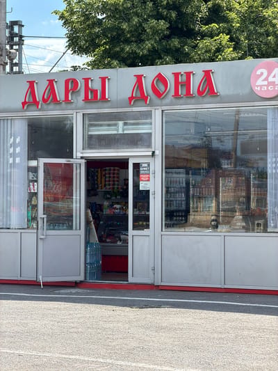 This is a medium shot of a retail kiosk with large glass windows and doors, located in Kamensk-Shakhtinsky, Russia. The kiosk has red lettering on its facade that reads "ДАРЫ ДОНА" (Dary Dona) and a circular sign with "24 часа" (24 hours) in pink. Inside the kiosk, shelves are stocked with various food items, including chips, drinks, and snacks. Several large bottles of water are stacked near the entrance. The ground in front of the kiosk is asphalt, with a white line marking a parking spot. Trees and overhead wires are visible in the background. The sky is blue with some clouds.