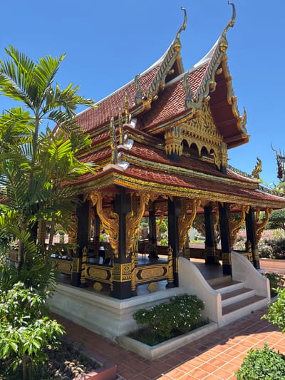 The image depicts a traditional Thai building located in the Bang Pu Subdistrict Municipality, Thailand. The building features a multi-tiered, red-tiled roof with elaborate gold ornamentation along the edges and gables. Black pillars adorned with golden carvings support the structure. A set of stairs leads up to a platform adjacent to the building, decorated with white concrete and flower beds with green plants. A brick-paved area surrounds the building's base. Lush green foliage, including palm trees, is visible in the foreground. The sky is a clear blue.