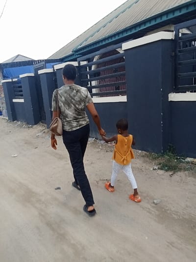 An adult female wearing a camouflage-patterned t-shirt, black pants, and sandals walks down a dirt road in Sapele, Nigeria (latitude: 5.862, longitude: 5.703). She is holding the hand of a young girl wearing a yellow top, white pants, and orange sandals. The woman carries a brown handbag over her right shoulder. Both are walking away from the camera, past a black and white wall with a metal gate to the right. The buildings in the background are covered with a light green roof. The ground is dirt and there are signs of tire tracks.