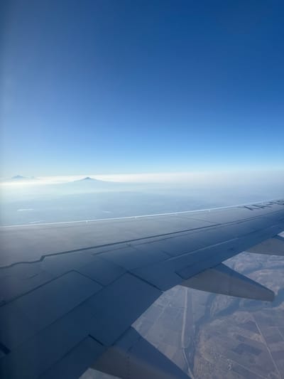 The image shows a view from an airplane window. The left side of the image is mostly a clear blue sky. Towards the center, there is a hazy horizon line with what appears to be a mountain in the distance, possibly shrouded in clouds or mist. The right side of the image is dominated by the wing of the airplane, which is grey in color. Below the wing, land is visible, showing a pattern that might be agricultural fields or a rural landscape. The image does not contain sufficient information to determine the specific location within North Miami Beach, United States of America. There is no depiction of people, specific subjects, objects, actions, or interactions visible in the photograph that would relate to the specified location.