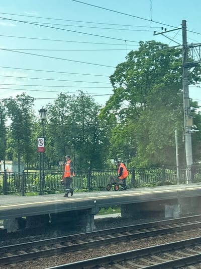 This image depicts a scene on a train platform in Lyuban, Russia, under a bright, slightly overcast sky with visible power lines crisscrossing the frame. In the foreground, train tracks are visible, indicating an active railway line. The middle ground features a train platform with two individuals in high-visibility orange vests and dark pants, suggesting they are likely maintenance or track workers.

The first individual, positioned on the left side of the platform, is walking and appears to be carrying an orange object. They are facing towards the right. The second individual, on the right side of the platform, is seated on a two-wheeled cart, also wearing an orange vest and dark pants. They are actively engaged with the cart, possibly in the process of moving it or preparing to.

Behind the platform, a black metal fence delineates the edge of the platform area. Beyond the fence, lush green trees dominate the background, with a street lamp visible on the left, featuring a "no entry" symbol with a red circle and diagonal line over a red symbol. The overall environment appears to be an outdoor railway station, likely in a more suburban or semi-rural setting given the dense foliage.