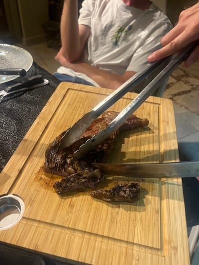 The image depicts a meal preparation scene taking place in Ganei Tikva, Israel, potentially in an outdoor or semi-outdoor setting. A large, seared cut of meat, exhibiting a dark, caramelized exterior and a reddish-pink interior, rests on a light-colored bamboo cutting board. A hand, visible on the right, uses silver metal tongs to stabilize the meat while another hand, also on the right, employs a silver-bladed knife to slice it. Multiple irregular slices of the meat are already separated on the cutting board, accompanied by visible meat juices. The cutting board features a perimeter groove and a circular metal insert. To the left, a section of a dining table is visible, draped with a dark, patterned tablecloth. On this table, a clear, textured glass plate and a set of silver cutlery (knife, fork, spoon) are present. In the background, a person with light skin is seated, wearing a white short-sleeved t-shirt adorned with a green and blue graphic. Their right arm is bent, resting. The floor in the right background consists of light-colored, irregularly shaped paving stones, consistent with an outdoor patio. The scene is illuminated by ambient light.