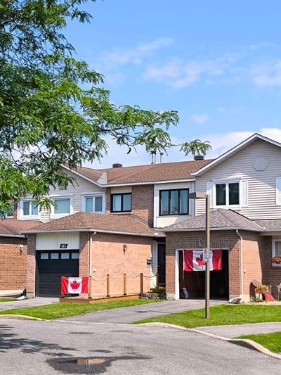 This media file captures a bright, sunny day in a suburban neighborhood in Ottawa, Canada. The scene features a row of attached brick houses with beige siding and brown roofs, indicative of a residential area. A large, leafy green tree partially obscures the upper left portion of the frame, suggesting it is summer or late spring. The sky is a vibrant blue with scattered white clouds, confirming fair weather.
The primary focus is on the houses, with Canadian flags prominently displayed. One flag hangs in the open garage of a house with the number "143" above the garage door. Another flag is visible on a small wooden deck in front of the same garage. The flags are a clear indicator of Canadian patriotism.
The houses are typical of North American suburban architecture, with multiple windows, some featuring white shutters. The landscaping includes neatly mown green lawns and paved driveways. A lamppost stands in front of one of the garages, with a circular sign attached, though the text on the sign is too small to be legible. The overall impression is one of a peaceful, well-maintained residential street on a clear, pleasant day, likely during a national celebration or holiday given the prominent display of Canadian flags. No people are visible in the frame, but the presence of flags suggests community pride and potentially a gathering or celebration, even if not explicitly shown.