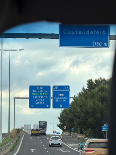 The image captures a view from inside a vehicle looking forward on a highway in El Prat de Llobregat, Spain. A large blue overhead sign indicates "Castelldefels 150 m" to the right. Below this, a series of blue directional signs are visible. The left sign, labeled "B-22," directs to "Barcelona," "Ronda de Dalt," "Tarragona," "Lleida," and "Girona" with downward-pointing arrows. The right sign, labeled "2A" and "C-31," directs to "Castelldefels" and "platges" with a downward-pointing arrow.

In the foreground, the highway has white lane markings and arrows. Several vehicles are on the road: a yellow minivan, a black car, a white car, and the rear of a gold SUV in the right lane. An Amazon truck with "There's more to Prime. A truckload more." branding is also present. To the left, a lamppost and a speed limit sign indicating "80" are visible. To the right, a dense line of green trees lines the highway. The sky is overcast with scattered clouds.