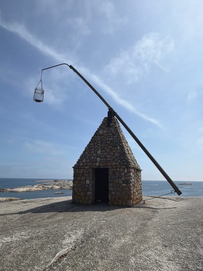 The image shows a small, stone structure with a conical roof, resembling a hut. A long, dark pole extends from the roof, with a metal cage-like object suspended from its end by a chain. The structure is situated on a rocky, flat surface overlooking the sea. In the background, there are islands and the blue ocean under a bright sky with wispy clouds. This is the "Vardehuset" on Verdens Ende, Tjøme, Norway. There are no people visible in the frame.