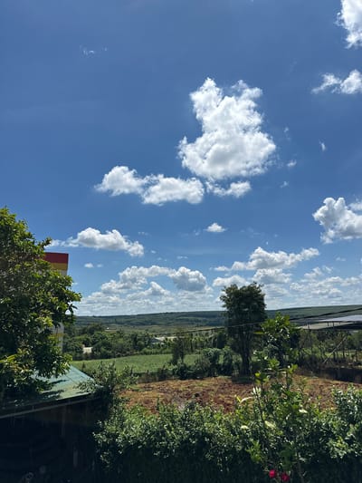 A landscape view in Ia Hru, Vietnam, features a vibrant blue sky filled with scattered white cumulus clouds. In the foreground, lush green trees and foliage partially obscure a building with a colorful facade of red and yellow bands. Beyond the immediate vegetation, a field of tilled brown earth is visible, leading to a more distant landscape of green hills and trees. A structure with a dark roof, possibly a greenhouse or shelter, with visible supports, is situated on the right side of the frame, partially covered by more greenery. The scene depicts a rural or semi-rural setting under a bright, clear sky.