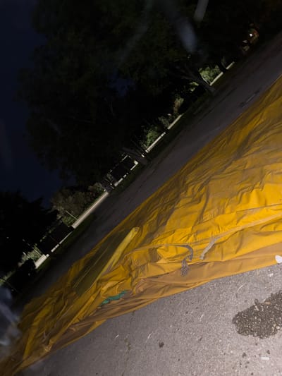 The image depicts a close-up view of a wrinkled yellow tarp, possibly covering an object or area. Adjacent to the tarp is a section of asphalt, likely a road or paved surface. Beyond the tarp, a dark, shadowed area contains trees and vegetation, suggesting a park or wooded area. The location is inferred to be Topoli, Bulgaria. The scene appears to be at night or in low-light conditions. No people are visible.
