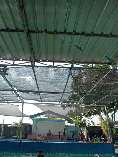 The image shows an outdoor swimming pool area at what appears to be a recreational facility. A metal roof structure with some sections covered by black mesh netting provides shade. Small, colorful pennant flags are strung across the underside of the roof. Several people are visible around the pool. In the foreground, a young male is partially submerged in the pool water. On the pool deck, to the left, a person sits on a bench under a yellow umbrella. Further to the right, several individuals are gathered, some standing and some sitting on a raised platform. A ladder leans against a wall behind them. A sign with text in Indonesian is mounted on the wall, indicating rules for the swimming pool. The environment suggests a sunny day, with trees and vegetation visible in the background. The presence of the swimming pool, the recreational setup, and the sign with local language information points to this location being a public or semi-public pool within a community, potentially Kemamang, Indonesia, given the user prompt.
