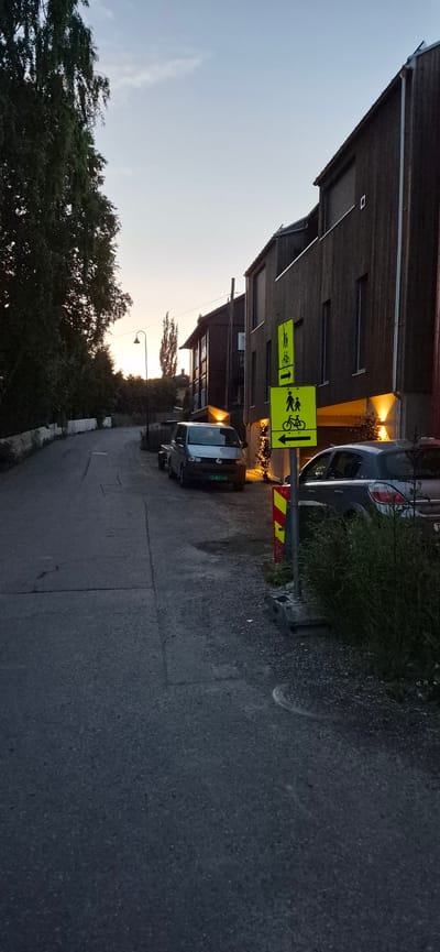 The image depicts a street scene in Lillehammer, Norway, during what appears to be dusk or early evening. A paved road curves to the left, lined with dark-colored wooden buildings on the right and trees and a stone wall on the left. Two vehicles are visible: a silver van parked on the right side of the road and a dark-colored hatchback parked further back. A street lamp is present in the middle distance. Two yellow traffic signs with pictograms indicating shared pedestrian and bicycle paths are prominently displayed on the right side of the road, with directional arrows. The sky is a pale blue with some cloud cover, and the ambient light suggests low sun or twilight. The overall impression is one of a quiet, residential street.