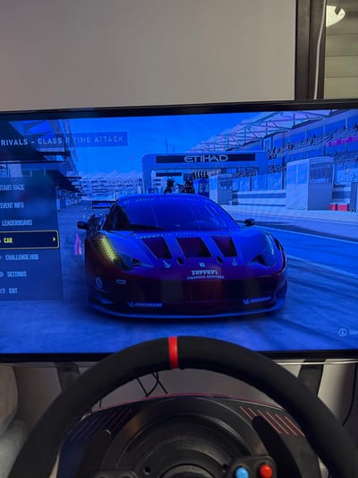 A screen displays a racing game menu, featuring "RIVALS - CLASS R TIME ATTACK". Selectable options include "START RACE", "EVENT INFO", "LEADERBOARD", "CAR", "CHALLENGE HUB", "SETTINGS", and "EXIT". A race car, adorned with "Ferrari FINANCIAL SERVICES" and "MICHELIN" logos, sits on the starting grid at the "ETIHAD" circuit. A gaming steering wheel is positioned in the foreground. A "Select" icon is visible at the top right.