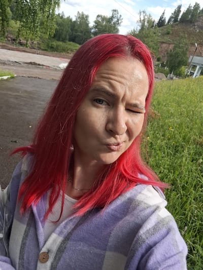 A woman with bright red hair is pictured in a close-up selfie taken outdoors. She is winking and puckering her lips, with her eyebrows furrowed. She is wearing a white tank top and a purple, grey, and white plaid shirt-jacket with a wooden button. The background shows a grassy area with scattered yellow flowers, a dirt road with some construction or landscaping materials, and a wooded hillside with some structures on it. The image appears to be taken in the city of Sim, Russia.