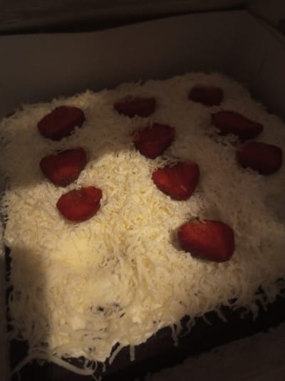 A rectangular cake is observed inside a light-colored, possibly cardboard, box in B. Nam Ngao, Laos. The cake features a dark base, likely chocolate, entirely covered by a thick layer of finely shredded white cheese. Ten whole, red strawberries are positioned on top of the cheese layer, distributed across the cake's surface. The strawberries appear ripe and have their green caps removed. The scene is dimly lit, with soft shadows visible within the confines of the box. No individuals are present.