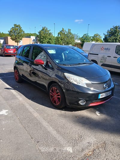 A black Nissan Note with red accents on the mirrors and lower bumper is parked in an outdoor paved lot. The car has red rims on its wheels. A red car is parked behind it. A white Dalkia EDF van is visible in the background. The sky is blue and clear, and trees line the perimeter of the lot. The scene appears to be a parking lot in Charleville-Mézières, France, given the French license plate on the Nissan Note, which reads "HH-776-FD."