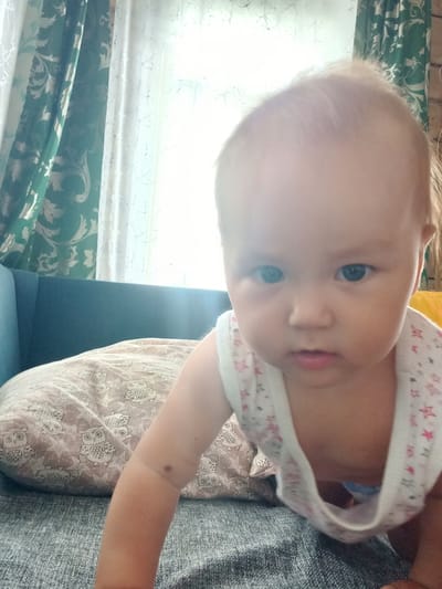 In the city of Идрисово, Russia, a close-up, eye-level, indoor shot captures a baby crawling on a gray, textured surface, possibly a couch. The baby, facing the camera, has fair skin, light brown hair, and dark eyes. They are wearing a white tank top with pink and red star patterns. To the left of the baby is a teal couch, and on it rests a light brown pillow with a repeating owl pattern. Behind the couch, green and white curtains are visible. The lighting suggests it is daytime, with bright light streaming in from a window behind the baby.