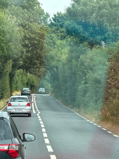The image depicts a road scene in the city of New Ross Municipal District, Ireland. A grey Volkswagen hatchback is in the foreground on the left, facing away from the camera, on a paved road lined with white dashed lines on both sides. Ahead of this vehicle, also on the road, is another grey hatchback, and further ahead, a dark-colored hatchback. The road is bordered by dense green trees and hedges on both sides, forming a tunnel-like effect. The sky is overcast and a faint glare is visible on the right side of the image. There are no people visible in the frame.