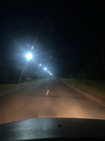 The image depicts a dark, rural road at night, illuminated by streetlights. The perspective is from inside a vehicle, looking forward. The road is paved and appears to have a single white line marking the center. On either side of the road, there are grassy areas and trees. Several tall streetlights line the left side of the road, their bright lights creating glare and lens flares within the image. The overall atmosphere is quiet and deserted, typical of a less populated area at night. The location is identified as Buchatsinski Sel'ski Savet, Belarus.
