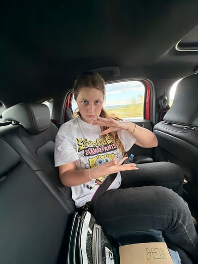 The image depicts a young woman sitting in the back seat of a car. She wears a white t-shirt with a "Spongebob Squarepants" graphic, black jeans, and bracelets on both wrists. Her hair is light brown. She is making hand gestures towards the camera. A cardboard box with the word "GAIA" is visible near her. A black and white handbag is next to her on the seat. The background shows a landscape through the car window, including fields and sky. The scene is presumably not in Kermen, Bulgaria because there is no evidence of this place in the picture.