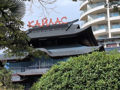 The image shows a building with a distinctive dark, multi-tiered, curved roof, reminiscent of traditional East Asian architecture. Red Cyrillic lettering spells out "КАЙЛАС" (Kaylas) across the top of the structure. This building is partially obscured by lush green foliage in the foreground and on the left side. In the background, a modern, white, multi-story building with curved balconies and glass railings stands against a pale sky. The overall scene suggests a location where modern and traditional architectural styles coexist, possibly a resort or a commercial establishment in a temperate climate, given the presence of greenery and the style of the buildings. This establishment is located in Guzeripl', Russia.