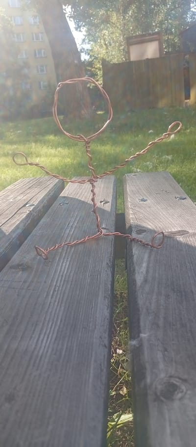 A wire figure, resembling a stick figure, is positioned on a weathered wooden park bench. The figure is made of copper-colored wire and is bent to appear as if it is sitting with its arms outstretched. The background shows a blurry residential building, a large tree, and a wooden fence, consistent with a park or outdoor public space within a city. The scene is illuminated by bright sunlight, creating lens flare effects. The exact location within Karviná, Czechia, is not discernible from the image alone.