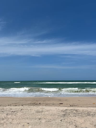The image shows a beach scene in Velamar, Mexico. The left side of the image is dominated by a clear blue sky with some wispy clouds. To the right of the sky is a strip of ocean with waves breaking on the shore. The rightmost section of the image is the sandy beach, with varying shades of tan and brown, suggesting different moisture levels and textures of sand. An orange object is seen on the beach. No people are visible in the image.