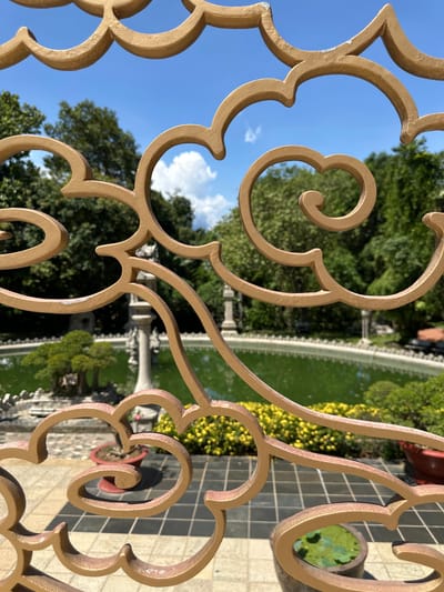 A close-up shot of an ornate, gold-colored metal fence partially obscures a view of a park in Dĩ An, Vietnam. The fence's design incorporates curls and floral motifs. Through the fence, the background reveals a rectangular pond or water feature, a stone fountain, manicured bushes, trees, a stone-tiled pathway or patio. In the foreground, a potted plant with green leaves is partially visible. The sky is blue with white clouds visible through the fence.