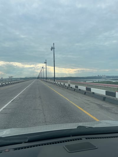 An eye-level, forward-facing, point-of-view shot from inside a vehicle on a bridge shows an overcast sky with sunbeams breaking through the clouds. The bridge stretches into the distance, lined with ornate streetlights and a distinctive white, black, and red railing. A river and a distant cityscape are visible on the right side of the frame. The road has clear lane markings, a white line dividing lanes in the foreground and a yellow line further ahead. The vehicle's dashboard is visible at the bottom of the frame.