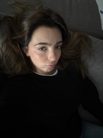 This image, taken in Nogueira e Silva Escura, Portugal, depicts a single female subject positioned horizontally in an indoor setting. The individual possesses long, dark brown hair, styled to be spread around her head. She has brown eyes, a fair complexion with a visible rosy tint on her cheeks, and dark eyebrows. Small, reflective earrings are visible in both ears. The subject is wearing a black long-sleeved top, with a white collar visible underneath at the neckline. A segment of blue fabric, consistent with denim, is visible in the lower right portion of the frame. Her gaze is directed forward, toward the camera, and her lips are slightly parted, forming a subtle pout. The background consists of a plain, dark grey, textured upholstered surface, indicating a piece of furniture such as a sofa or bed.