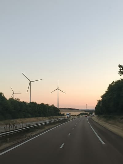 The image shows a view from a highway in Joux-la-Ville, France, at dusk. In the distance, several wind turbines are visible, silhouetted against a pale orange and blue sky. The highway curves to the right, with white lines marking the lanes. On the left side of the road, there is a line of trees and vegetation. On the right side, more trees and a slightly elevated embankment are visible. A few vehicles, including a truck and a car, are driving on the highway. The overall scene suggests a journey through a rural landscape during twilight.