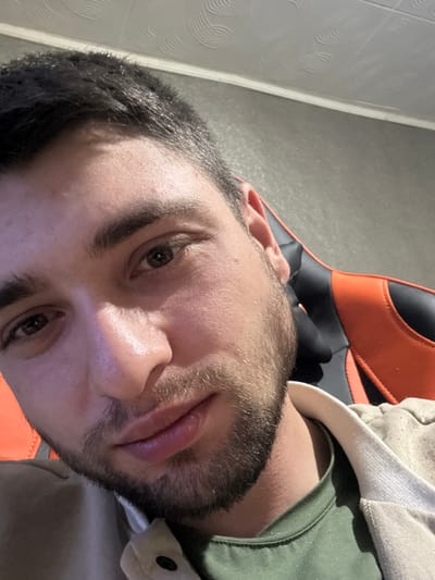 The image presents a close-up portrait of a male individual, appearing to be a young adult. He has short, dark hair and a short beard or stubble. The individual is looking directly into the camera. He is wearing a dark green t-shirt layered under a light beige or off-white collared jacket or shirt. Behind him, to the right, a portion of a black and orange gaming chair is visible. The background consists of a plain light-colored wall, and the upper section of the image displays a white ceiling with a distinct, repeating circular or spiral pattern. The scene is indoors. The specific location within Profesor Ishirkovo, Bulgaria, cannot be determined from the visual information provided.