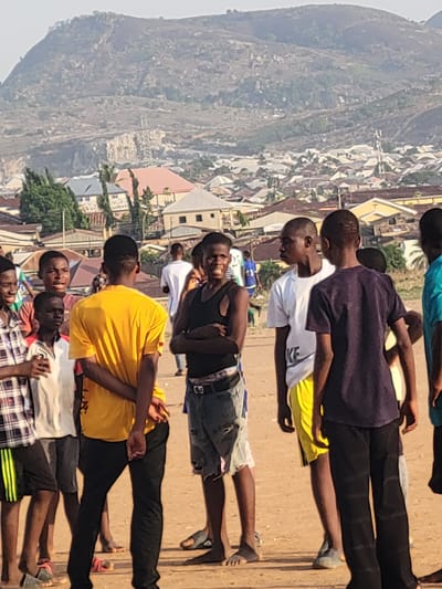A group of people, primarily young men, are arranged in rows on a dirt surface. Some are lying down while others are seated. The setting appears to be outdoors with a blurred background of buildings and a mountain visible in the distance. The location is Ibo, Nigeria, based on the urban landscape and geographical features in the backdrop. The specific activities or interactions are not readily apparent. The lighting suggests it's daytime.