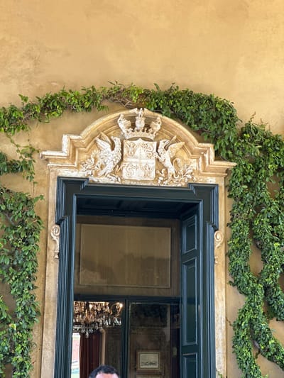 This image captures a detail of an exterior wall in Tremezzina, Italy. The wall is a warm, sandy yellow, textured and slightly weathered. A dark green, ornate window frame with detailed moldings is set into the wall. To the left of the window, a circular, sculpted emblem adorns the wall. This emblem features intricate white relief carvings, including what appears to be a heraldic crest with a book at its center, flanked by mythical creatures that resemble griffins or winged lions. The emblem is surrounded by a decorative, layered frame.
Green ivy or a similar vine with small, dense leaves cascades over and around the window and the sculpted emblem, adding a touch of nature to the architecture. The ivy is lush and vibrant, suggesting a pleasant, possibly mild climate.
The window itself is reflective, showing a distorted view of an interior space. We can discern reflections of what looks like a chandelier with multiple crystals, and possibly framed artwork on a wall within. The light suggests it might be daytime, but the interior reflections are somewhat dim, perhaps indicating the room is lit by artificial light or is in shadow.
The image is a close-up shot, focusing on the architectural details and the interplay of the vine with the stonework and window. There is no visible text in the image. The overall impression is one of historical charm and rustic elegance, typical of Italian villas or buildings in scenic locations.