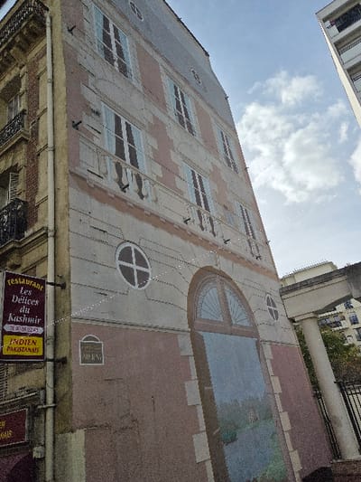 The image depicts a tall, narrow building facade featuring a trompe l'oeil mural, located at 5 Rue Henri Barbusse in Levallois-Perret, France. The mural simulates a classic architectural style, showing multiple floors with faux windows featuring light blue shutters, intricate sills, and balcony railings painted in black. A large, arched, multi-paneled window is depicted at the bottom, opening onto a painted landscape scene with a body of water and a distant building. Several circular faux windows are also incorporated into the mural's design.
To the immediate left, a real, more traditionally styled building with ornate stone detailing and actual iron balconies is partially visible. A white vertical pipe runs along the corner separating the two structures. Attached to this traditional building is a black rectangular sign for "Restaurant Les Délices du Kashmir," displaying a phone number "01 40 88 22 43" and "INDIEN PAKISTANAIS" below it. Below this, another sign fragment is visible. A smaller, rounded rectangular street sign on the mural wall reads "Rue M HENRI."
The sky overhead is partly cloudy blue, and a modern high-rise building is partially visible in the upper right background. No people are present in the frame. The overall perspective is upward-looking, emphasizing the height of the mural.