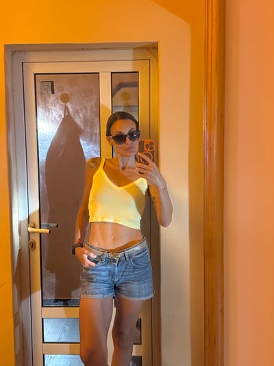 A woman takes a selfie in front of a doorway. She is wearing a yellow crop top, denim shorts, sunglasses, and a watch. Her phone is visible in her right hand. She has a tattoo on her left arm. The doorway appears to be made of glass and metal, and it is illuminated by warm, orange light. The background is a peach-colored wall with a vertical wooden post. The scene does not provide any specific indicators to pinpoint a location within the city of Pleven, Bulgaria.