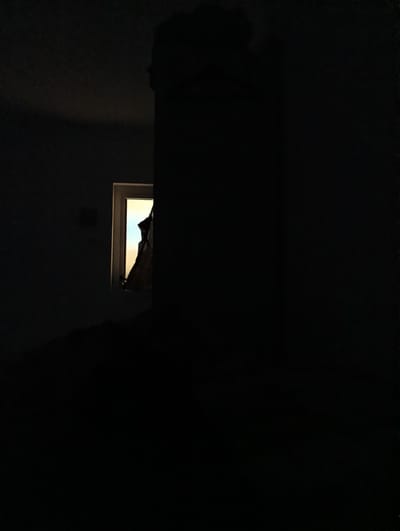 A dimly lit room is depicted with a small window on the left side. The window appears to be open, and a piece of fabric, possibly a curtain or garment, is hanging from it, obscuring part of the view outside. The fabric is dark and appears to have some texture. The light coming through the window is a pale yellow or orange, suggesting it might be daylight or artificial light from outside. The rest of the room is in deep shadow, with a large, dark, solid object occupying the right side of the frame. The overall impression is one of darkness and obscurity, with the window providing a small, illuminated focal point. The scene is devoid of any discernible activity or interaction.