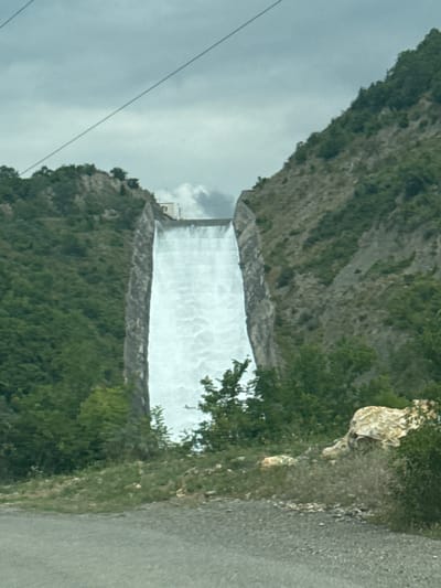 The image shows a massive concrete dam with water flowing over its spillway, creating a wide, frothy waterfall. The dam is situated within a steep, mountainous ravine, with dense green vegetation covering the slopes on either side. A building, possibly part of the dam's infrastructure, is visible at the top of the spillway on the left side. The sky is overcast with gray clouds, and a power line stretches diagonally across the upper portion of the frame. In the foreground, a gravel road or path is visible, with some rocky outcrops and sparse vegetation. The scene appears to be the Zhinvali Dam in Georgia, a prominent landmark known for its hydroelectric power generation. There are no people or animals visible in the image.