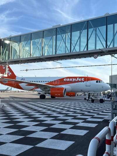 The image displays an EasyJet Airbus A320 aircraft, registered OE-LKF, parked on the tarmac at Nice Côte d'Azur Airport (NCE) in Saint-Laurent-du-Var, France. The aircraft, featuring a white fuselage with an orange tail and the "easyJet" logo in orange text, is connected to a transparent, glass-paneled jet bridge. The jet bridge exhibits structural cross-bracing visible through its panels. Below the jet bridge, a red circular sign with "4 m" in white text indicates a maximum height clearance. The tarmac in the foreground is marked with a distinctive black and white checkered pattern. Several pieces of ground support equipment are present near the aircraft; a white vehicle is positioned near the nose landing gear, and another partially visible yellow-and-white vehicle is closer to the nose. White and red striped barriers are visible in the lower right foreground. The background reveals a clear blue sky with some wispy white clouds and parts of other aircraft in the distance. No individuals are visible within the frame.