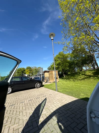 The image shows an outdoor scene, likely in Wassenberg, Germany, due to the prompt's indication. A car, seemingly a black sedan, is parked on a brick-paved surface, possibly a driveway or parking area. The car door is open, partially obscuring the view. A lamppost stands nearby, casting a shadow on the paving. In the background, there is a small wooden shed and a grassy area with trees. The sky is blue with some scattered clouds, suggesting a clear day.