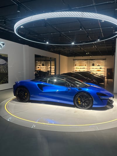 The image shows a blue McLaren Artura sports car displayed on a circular platform with embedded lighting in a showroom. Several other vehicles of varying colors (black, grey) are visible behind the Artura. The location is in Glattbrugg, Switzerland, as evidenced by the McLaren store's presence in that city. The showroom features a modern design with a high ceiling and a large circular lighting fixture. The walls are adorned with displays, suggesting a retail environment for McLaren merchandise. No people are visible in the frame.