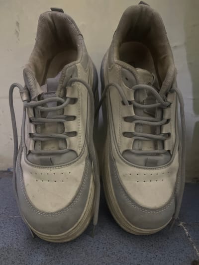 The image displays a pair of grey and white athletic shoes placed on a speckled blue and grey floor. The shoes are positioned one above the other, with the top shoe slightly overlapping the bottom one. Both shoes are laced with grey laces that are threaded through loops on the top of the shoe. The shoes appear to be well-worn, with some scuff marks and dirt visible on the white and grey sections. The interior of the shoes, visible in the opening, also shows signs of wear. The lighting in the image suggests it was taken indoors during the daytime. There is no text visible in the image. The scene is a simple, everyday setting, likely a home or a casual indoor space.