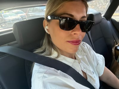 A woman wearing sunglasses and a white shirt is sitting in the backseat of a car. She has blonde hair and is wearing white earbuds. She is looking at the camera. The car appears to be in motion, and the background shows other cars and a stone wall, suggesting an urban or suburban environment. The image's metadata indicates the location as Nanterre, France.